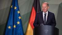 Olaf Scholz steht an einem Rednerpult, dahinter die Flagge Deutschlands und der EU