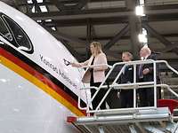 Taufe A350 Konrad Adenauer Eine Frau und zwei Männer stehen in einer Halle auf einer Plattform neben einem weißen Flugzeug