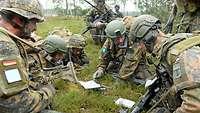 Eine Gruppe von Soldaten liegt im Gras in einem Kreis und bespricht ihre Lage. Weitere Soldaten befinden sich im Hintergrund..