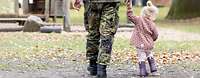 Ein Soldat geht mit seiner kleinen Tochter über eine mit Laub bedeckte Wiese. Beide sind nur von hinten zu sehen.