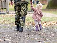 Auf dem weg zur Kita Ein Soldat geht mit seiner kleinen Tochter über eine mit Laub bedeckte Wiese. Beide sind nur von hinten zu sehen.