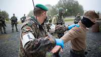 Ein Soldat kontrolliert eine Person. Im Halbkreis stehen weitere Soldaten, die diesen Vorgang beobachten.