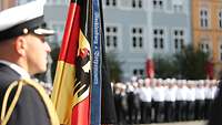 Ein Marinesoldat in Uniform steht neben einer Flagge der Bundesrepublik Deutschland angetreten vor einer Häuserfront.
