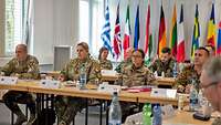 Soldiers from different nations sit at tables and listen to a lecture. Flags of different nations are in the background.