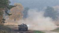 Ein Kampfpanzer mit einer roten Flagge steht schießend auf einer grünen Waldlichtung im Staub.