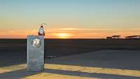 Eine Miniatur-Betonsperre mit einer Weihnachtsmannfigur steht im Flugbetriebsbereich – im Hintergrund der Sonnenuntergang 