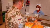 Ein Soldat steht mit Tablett und Teller an der Essensausgabe. Ein ziviler Mitarbeiter gibt Essen aus.