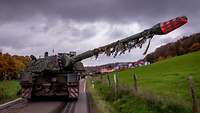 Auf einer Landstraße vor einem Dorf steht eine Panzerhaubitze mit erhöhtem Rohr.