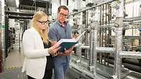 A woman and a man stand next to the technical installations of a building and inspect documents