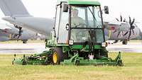 A man sits in the cab of a riding lawn mower and mows the lawn at an airfield; in the background, an Airbus A400M can be seen.