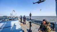 A man in uniform stands next to a microphone on the deck of a ship; in the background, a helicopter can be seen.