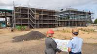 A woman in civilian clothing and a man in uniform look at a site plan; in the background, a construction site can be seen.