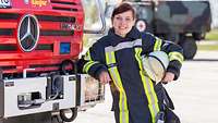 A woman in uniform holds a helmet in her hand and leans against a fire truck.