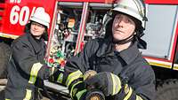 Two firefighters stand in front of a fire truck, one is holding a hose in his hand