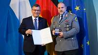Ein Soldat und ein Zivilist stehen mit einer Urkunde und einem Orden vor großen Flaggen.