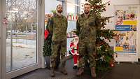 Zwei Soldaten stehen vor einem geschmückten Weihnachtsbaum. Daneben ein Plakat mit dem Spendenzweck, für den gesammelt wird.