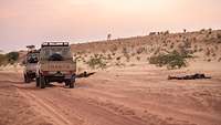 Zwei Pick-Ups halten auf einer Sandpiste. Die Insassen liegen im Sand und erwidern das Feuer.