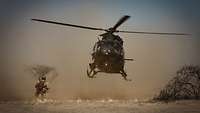 Ein Hubschrauber befindet sich im Landeanflug davor kniet ein Soldat im Savannensand