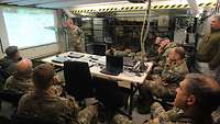 Ein Soldat am Whiteboard zeigt auf eine Karte, mehrere Soldaten sitzen um einen Tisch versammelt und hören zu 
