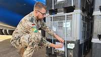 Ein Soldat in Wüstentarnuniform kniet am Boden und befestigt ein Schild an einer Kiste