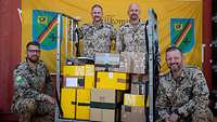 Gruppenfoto mit vier Soldaten der Feldpost, mittig ein Wagen mit Paketen, hinter ihnen ein Banner mit dem Wappen der Feldpost 
