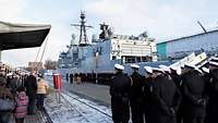 Auf einer Pier neben einem grauen Kriegsschiff sind Marinesoldaten in dunkelblauen Mänteln angetreten.