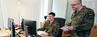Ein Soldat sitzt an einem Schreibtisch vor einem Computer, neben ihm steht ein anderer Soldat