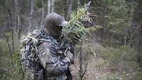 Ein Soldat, getarnt mit Ästen und spezieller Tarnkleidung bewegt sich lautlos durch den Wald.