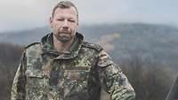 Ein Soldat im Dienstgrad Oberfeldwebel blickt in die Kamera. Im Hintergrund ist eine hügelige Landschaft.