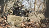 Ein Soldat liegt mit seiner Waffe auf dem Waldboden hinter seinem Rucksack. Im Vordergrund liegen Blätter und große Steine. 