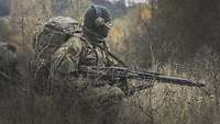 Zwischen hohem Gras und Dornenbüschen kniet ein Soldat mit Maschinengewehr MG3 im Anschlag
