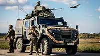 Ein gepanzertes Militärfahrzeug vom Typ Eagle V mit Soldaten. Im Hintergrund ist ein Kampfflugzeug in der Luft.
