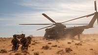 Zwei Soldaten in Wüstentarnuniform mit Helm warten vor einem Hubschrauber mit drehenden Rotoren