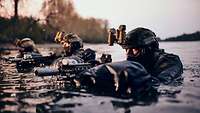 Drei Soldaten schwimmen mit dem Gewehr im Anschlag im Wasser im Morgenlicht.