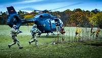 Luftgestützte Operation Zwei Soldaten stürmen mit dem Gewehr über eine Waldlichtung. Dahinter landet ein grauer Hubschrauber.