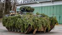 Ein mit grünen Ästen getarnter Panzer fährt auf einer Straße zwischen Hallen.