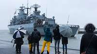 Familienangehörige verabschieden sich von dem Kriegsschiff „Mecklenburg-Vorpommern“. Das Wetter ist trüb und es regnet..