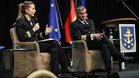 Eine Marineoffizierin und ein Admiral in dunkelblauer Uniform sitzen auf einer Bühne.