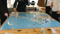 Dice and game pieces are spread out on a map, with four people pondering the right strategy