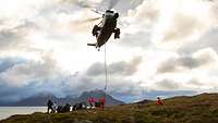 Ein Hubschrauber schwebt in der Luft und lässt per Seil Kisten runter. Mehrere Personen stehen unter ihm am Boden.