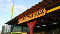 Ein gelbes Schild mit der schwarzen Aufschrift „Feldpostamt Sliač“ hängt unter einem Vordach