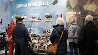 Mehrere Zivilisten betrachten in einem Besucherzentrum eine Ausstellungswand mit Bildern, Texten und Exponaten.