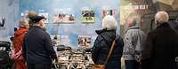 Mehrere Zivilisten betrachten in einem Besucherzentrum eine Ausstellungswand mit Bildern, Texten und Exponaten.