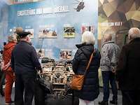 Öffentliche Ausstellung Mehrere Zivilisten betrachten in einem Besucherzentrum eine Ausstellungswand mit Bildern, Texten und Exponaten.