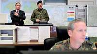 Links stehen ein Mann im Anzug und ein Soldat vor vielen Landkarten und Schreibtischen, vorn rechts ein Soldat vor einem Monitor