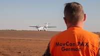 Ein Soldat mit einer Warnweste mit dem Aufdruck MovCon PAX German steht am Flughafen Gao, am Horizont eine Propellermaschine