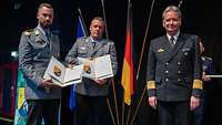 Ein Soldat übergibt zwei Soldaten jeweils eine Urkunde und einen Preis.