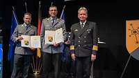 Ein Soldat übergibt zwei Soldaten jeweils eine Urkunde und einen Preis.