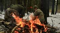 Drei Soldaten hacken im Wald Feuerholz. Im Vordergrund brennt ein Wärmefeuer.