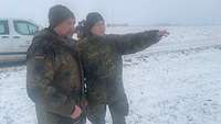 Zwei Soldaten stehen auf einem Feld. Eine Soldatin zeigt mit dem gehobenen linken Arm nach rechts
